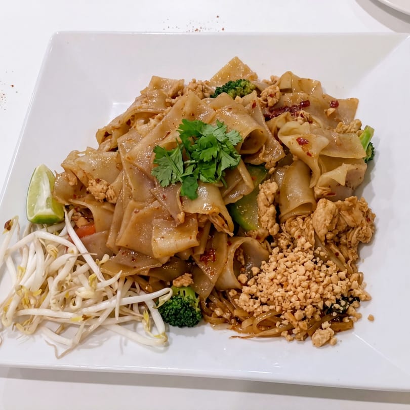 Pad See Ew with chicken, broccoli, bean sprouts, lime, and crushed peanuts on a white plate.