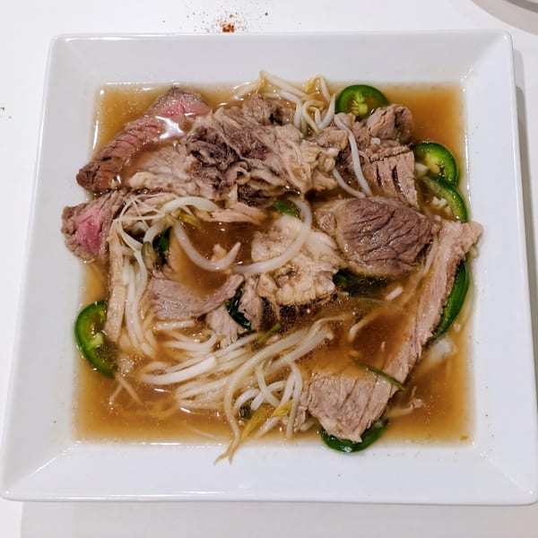 A square white bowl of pho with beef, noodles, bean sprouts, and sliced jalapeños in broth.