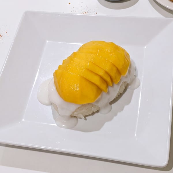 Mango sticky rice dessert with sliced mango and coconut milk on a white rectangular plate.