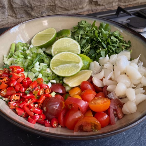A bowl filled with chopped red chilies, green onions, cherry tomatoes, pearl onions, cilantro, and lime wedges.