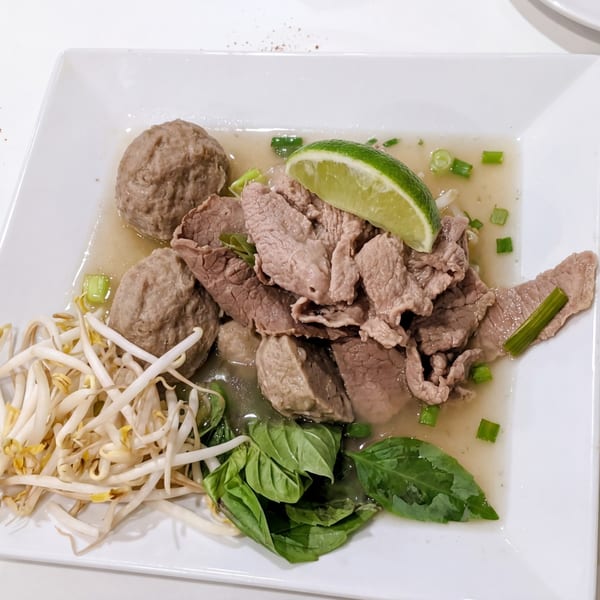 A white rectangular plate holds a bowl of pho with beef, meatballs, bean sprouts, basil, and lime.